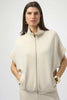 A woman with long dark hair models the Joseph Ribkoff Scuba Jersey Boxy Jacket 254145 in cream, featuring short sleeves and a stand collar. She accessorizes with gold hoops and bracelet, posing hands-in-pocket against a light background.