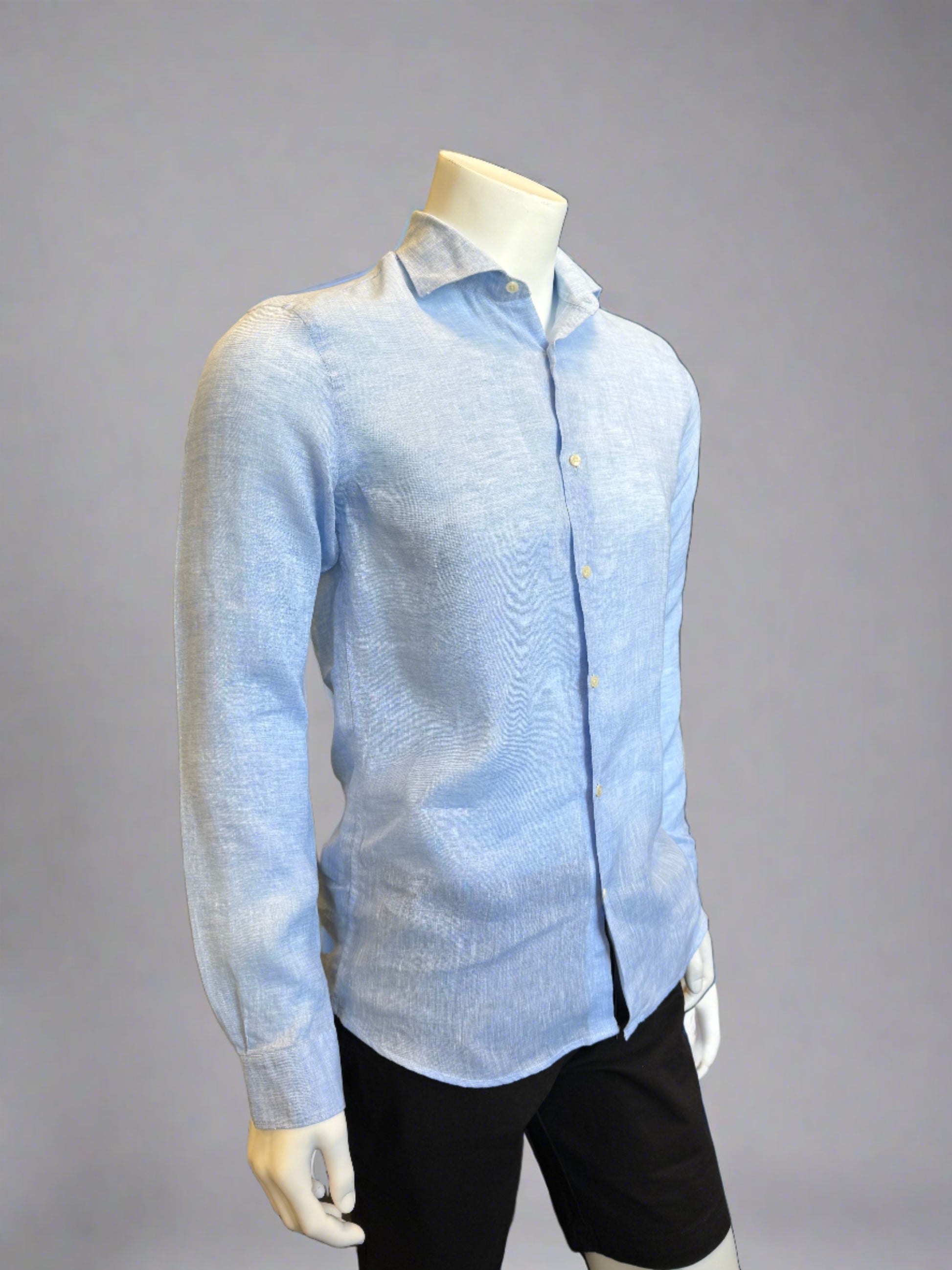 A headless mannequin showcases a Paul Miranda Linen Shirt Cielo in light blue. Crafted in Italy from 100% linen, the slim-fit shirt is adorned with front buttons and buttoned cuffs. The mannequin, also sporting black pants, stands against a plain gray background, highlighting the shirt as the main focus.