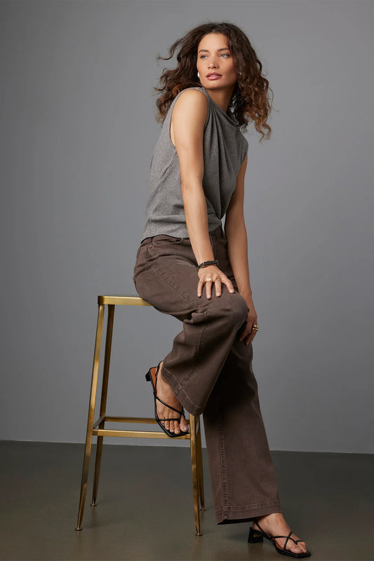 A woman with curly hair sits sideways on a gold stool against a gray backdrop, wearing a sleeveless gray top and Wit & Wisdom Sam High Rise Wide Leg jeans (P2652LEPE). She pairs them with black strappy heels and looks relaxed.