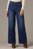 A person wearing Wit & Wisdom WHITNEY SKYRISE WIDE LEG JEANS in blue, paired with a cream long-sleeve top and brown heeled boots, stands against a plain background.