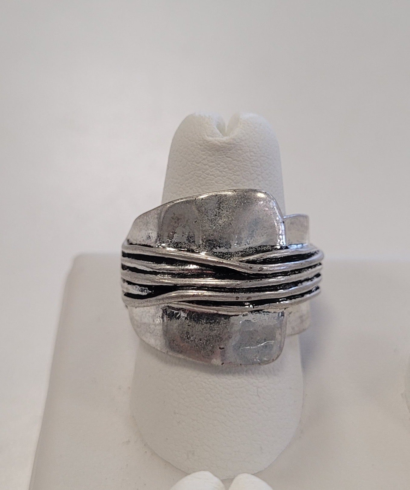 The Jenna-74 Adjustable Ring by Elise accessories features silver, asymmetrical overlapping layers and multiple thin horizontal bands at the center, shown on a white ring holder—an eye-catching statement piece.