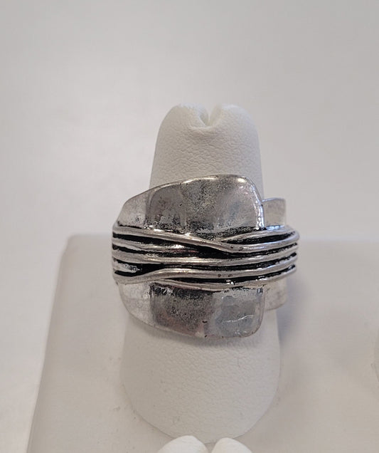 The Jenna-74 Adjustable Ring by Elise accessories features silver, asymmetrical overlapping layers and multiple thin horizontal bands at the center, shown on a white ring holder—an eye-catching statement piece.