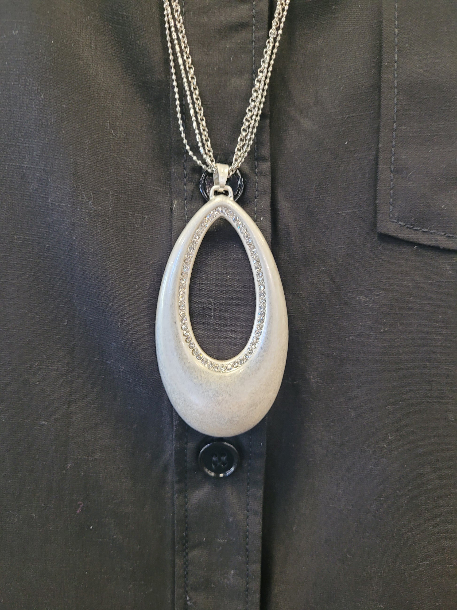 The Andy-23 Necklace by Elise accessories features a large, metallic teardrop pendant with a textured edge on an elegant two-strand chain, creating striking contrast against a black button-up shirt.