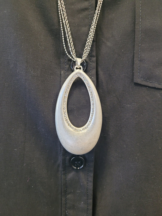The Andy-23 Necklace by Elise accessories features a large, metallic teardrop pendant with a textured edge on an elegant two-strand chain, creating striking contrast against a black button-up shirt.