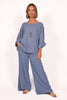 A person is wearing the Cotton Gauze Pants ITS70031 and a matching top by Catherine Lilywhite. The blue outfit, made in Italy, is loose-fitting. They accessorize with a long necklace with circular pendants and sandals, posing with one hand in a pocket against a plain white background.