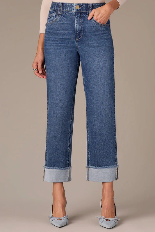 A person stands against a beige background, showing only their lower torso and legs, wearing Wit & Wisdom HIGH RISE CLEAN FINISH FRAY CUFF HEM PETITE BOYFRIEND JEANS and pointed denim shoes.