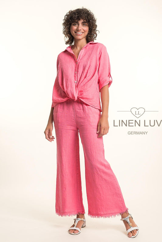 A woman in the LinenLuv French Linen Shirt TP1699-219 Geranio 44 and matching wide-leg pants with frayed hems, paired with white sandals, smiles against a light background. The Linen Luv Germany logo is visible.