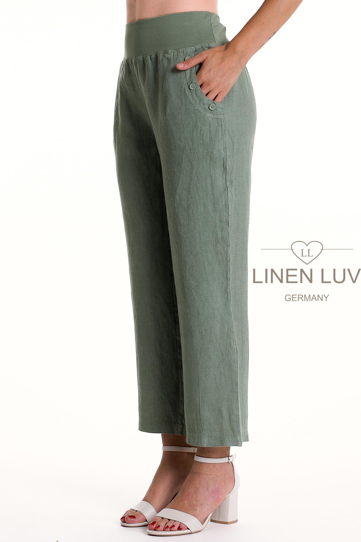 A model wears LinenLuv's French Linen Pant PT875-167 Sauge—sage green, high-waisted, wide-leg linen trousers with side buttons and pockets. Styled with white heeled sandals against a white background featuring the Linen Luv Germany logo.