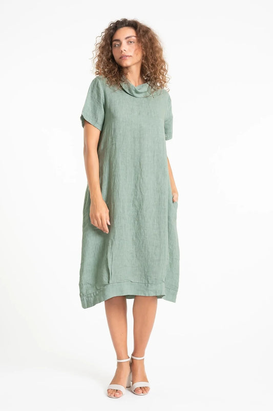 A curly-haired woman stands against a white background in a loose-fitting, knee-length French Linen Dress TP1557-167 Sage from LinenLuv. The eco-friendly European linen dress features short sleeves and a cowl neck. She pairs it with white strappy sandals, her hand casually in her pocket.