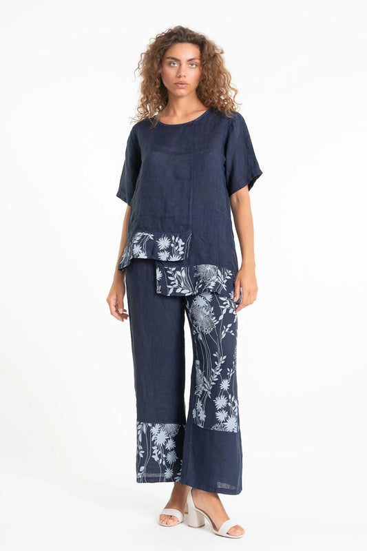 A woman wears LinenLuv's TP1605P-17 BLU/S, a French Linen Blouse in dark blue with white floral designs. The outfit includes a short-sleeve top and wide-leg pants. She stands against a plain white background, wearing light-colored, open-toe heels that reflect her eco-friendly style.