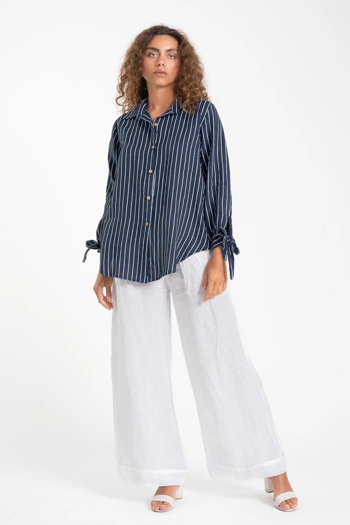 A person with curly hair models the French Linen Shirt TP1600-17 BLU/S by LinenLuv, a navy striped button-up with tied sleeves, paired with white wide-leg pants and open-toed shoes against a plain white background, their neutral expression complementing the eco-friendly outfit.