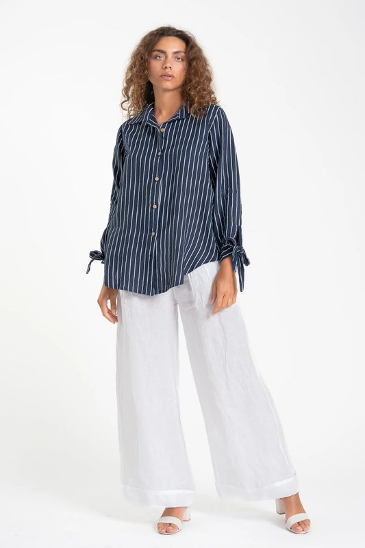 A person with curly hair models the French Linen Shirt TP1600-17 BLU/S by LinenLuv, a navy striped button-up with tied sleeves, paired with white wide-leg pants and open-toed shoes against a plain white background, their neutral expression complementing the eco-friendly outfit.