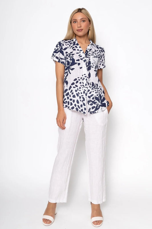 The individual is donning a French Linen Blouse TP1485-11 Bianco by LinenLuv, showcasing a navy and white floral pattern on eco-friendly fabric. Paired with white pants and sandals, they have long blonde hair against a plain white background.