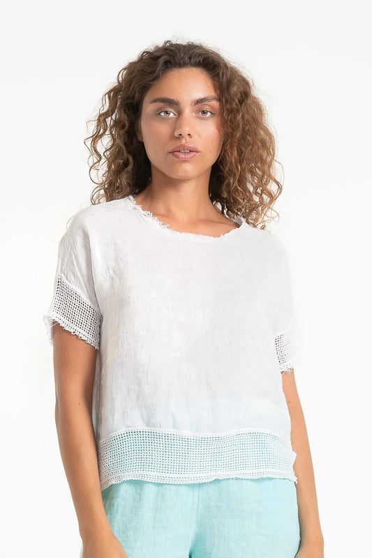 A person with curly hair is wearing the French Linen Top (TP1524-11 Bianco) by LinenLuv, featuring short sleeves and mesh trim, paired with light blue eco-friendly European linen pants. They stand against a plain white background.