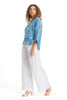 A curly-haired person wears the eco-friendly LinenLuv "French Linen Top TP1552P-185 Avio," featuring a blue shade with white patterns. They pair it with loose-fitting white pants and white heeled sandals, posing side-on against a plain white background.
