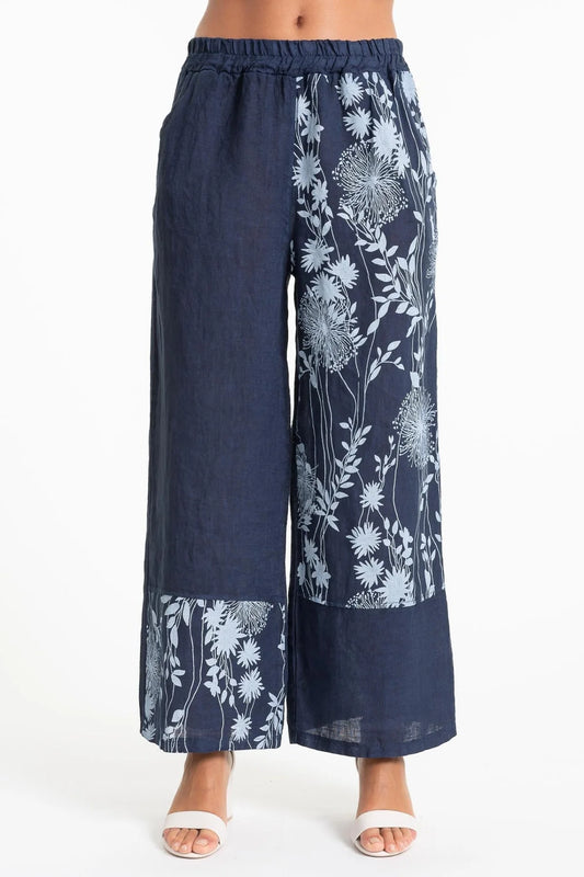 A person stands barefoot against a white backdrop, showcasing LinenLuv's French Linen Pant PT852-17 BLU/S. The navy blue wide-leg pants with light blue florals enhance their brown skin, exuding an effortlessly chic and eco-friendly vibe.