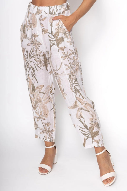 A model showcases eco-friendly LinenLuv's French Linen Pant PT832-11 Bianco, featuring a floral pattern in beige and green on white 100% European linen. With a relaxed fit, these pants are paired with white open-toe heels, as the model casually places a hand in the pocket.