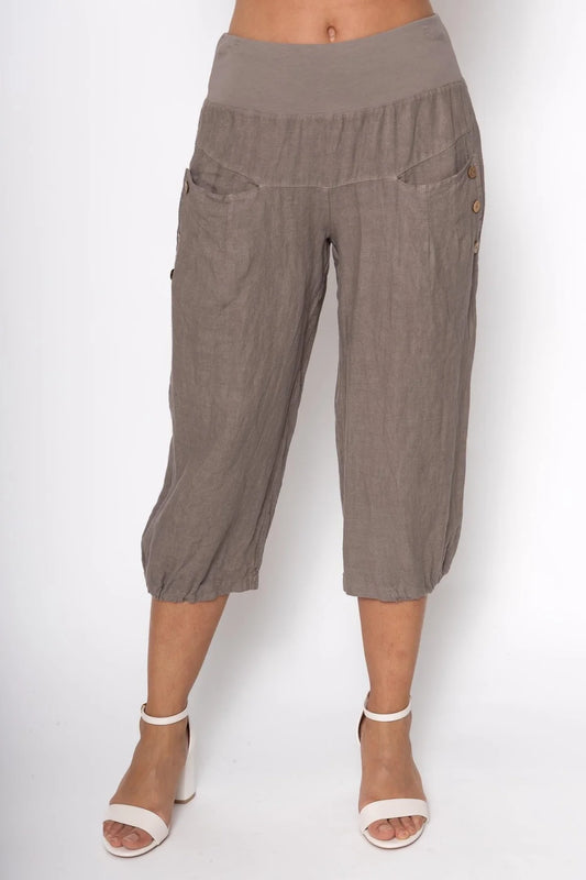 A person is wearing loose-fitting, cropped French Linen Capri PT836-40 by LinenLuv in light brown. The pants feature a wide waistband with two side buttons and are made from eco-friendly 100% European linen. They stand barefoot with white ankle-strap sandals against a plain white background.