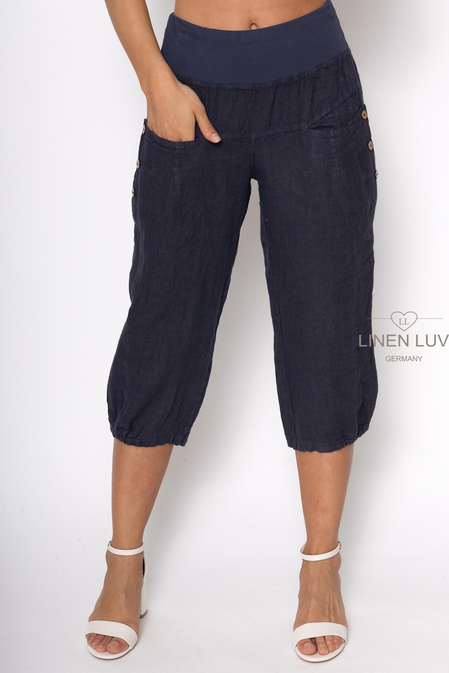 Someone is wearing dark blue French Linen Pant PT836-71 - Sabbia from LinenLuv with a wide waistband and side buttons, paired with white high-heeled sandals. Their left hand rests in a pocket, showcasing the brand's comfort and style on the right side.