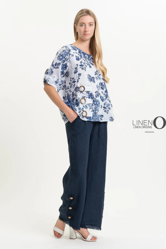 A woman elegantly poses sideways in the French Linen Blouse TP1519P-11 Bianco by LinenO, featuring a floral blue and white design with wide sleeves and button details, complemented by dark wide-leg pants with ankle buttons.