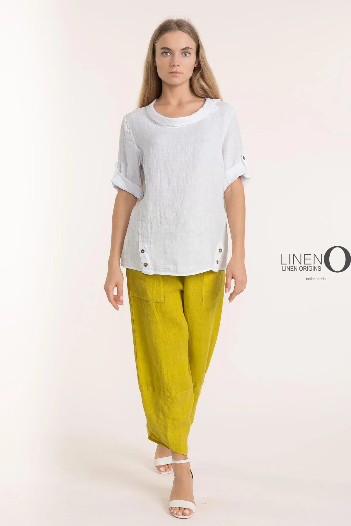 Dressed in LinenO's eco-friendly French Linen Blouse TP1340-11 Bianco, a person with long hair and rolled-up sleeves paired with yellow linen pants strides confidently in white open-toed sandals. The minimalist style stands out against a plain backdrop, emphasizing sustainable fashion's allure.