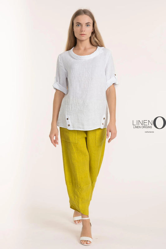 Dressed in LinenO's eco-friendly French Linen Blouse TP1340-11 Bianco, a person with long hair and rolled-up sleeves paired with yellow linen pants strides confidently in white open-toed sandals. The minimalist style stands out against a plain backdrop, emphasizing sustainable fashion's allure.