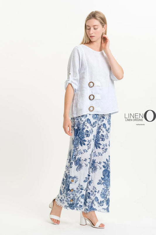A woman in a French Linen Blouse TP1519-11 Bianco from LinenO pairs it with floral blue and white wide-leg pants. Against a plain white background, she touches her hair while looking down. Text reads "LINEN ORIGINS.