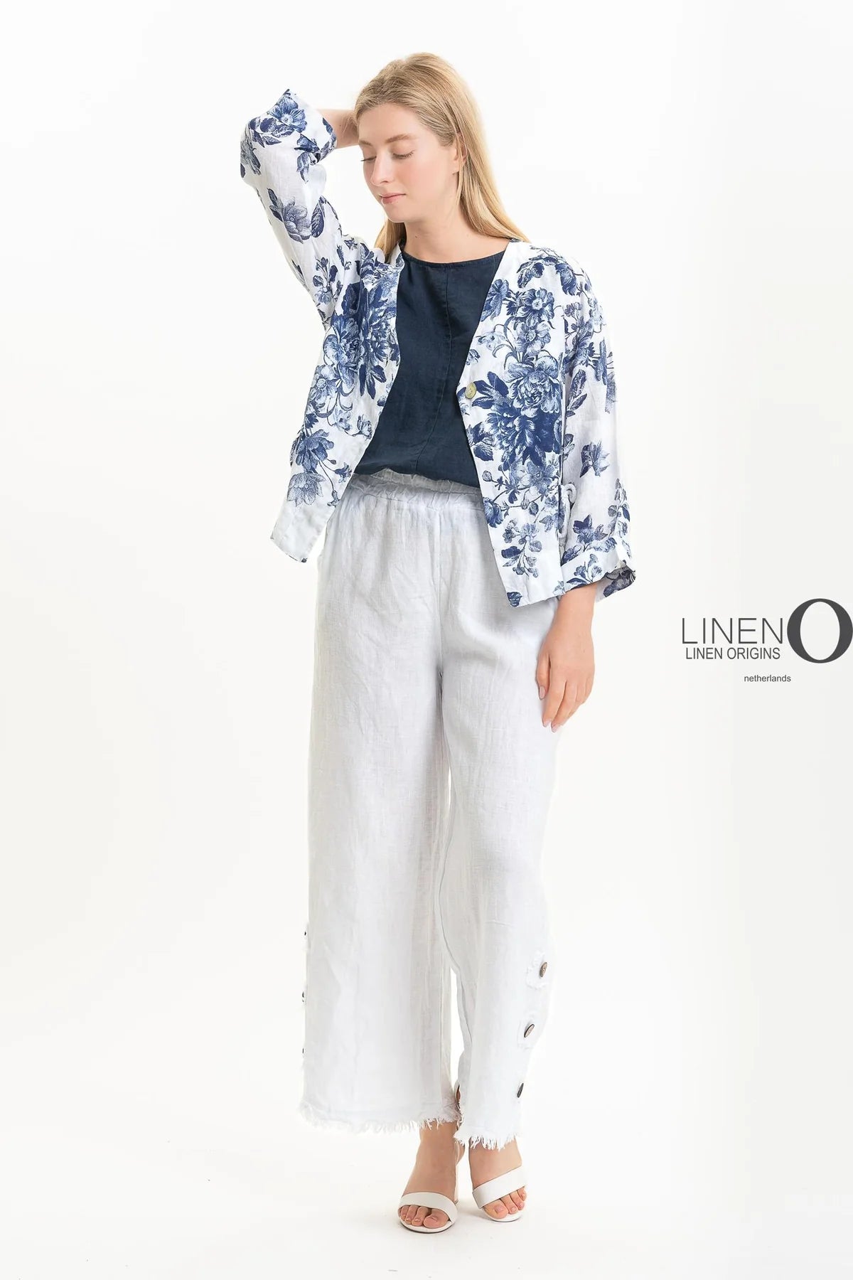 Against a white background, a person wears the French Linen Jacket TP1523P-11 Bianco by LinenO with a dark blue top, white wide-legged pants, and white sandals. Made from eco-friendly French linen; they stand relaxed with one hand on their forehead. The "LINEN ORIGINS" logo is visible on the right.