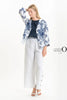 Against a white background, a person wears the French Linen Jacket TP1523P-11 Bianco by LinenO with a dark blue top, white wide-legged pants, and white sandals. Made from eco-friendly French linen; they stand relaxed with one hand on their forehead. The "LINEN ORIGINS" logo is visible on the right.
