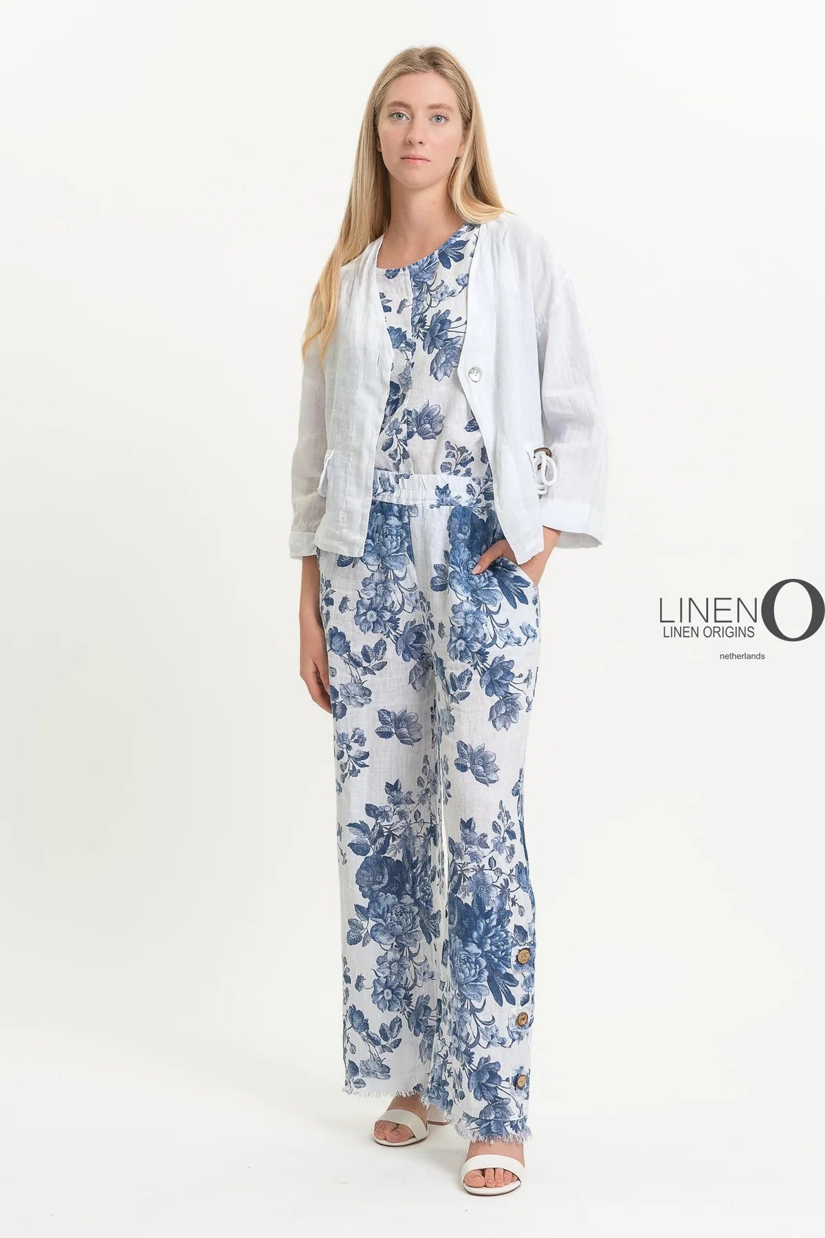 A woman stands against a plain background, wearing an eco-friendly white linen outfit with blue floral patterns. Her ensemble features the French Linen Jacket TP1523-11 Bianco by LinenO, paired with matching top and pants. White sandals complete the look, while "LinenO" appears nearby.