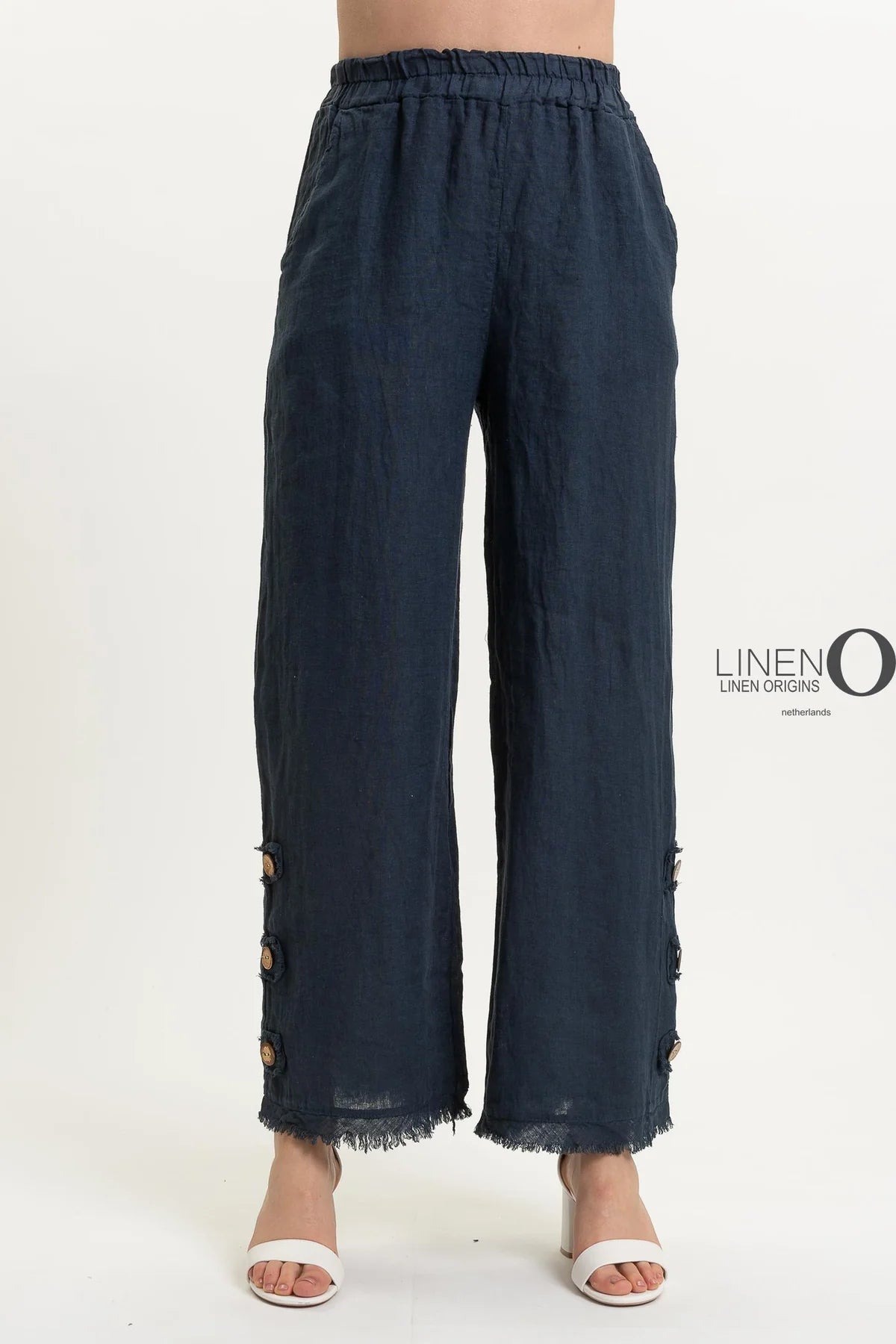 A person wears the stylish French Linen Pant PT839-75 Notte by LinenO, featuring wide-leg dark blue European linen with frayed hem and wooden buttons near the bottom. Paired with white open-toed heels on a crisp white background, it highlights eco-friendly fashion choices.