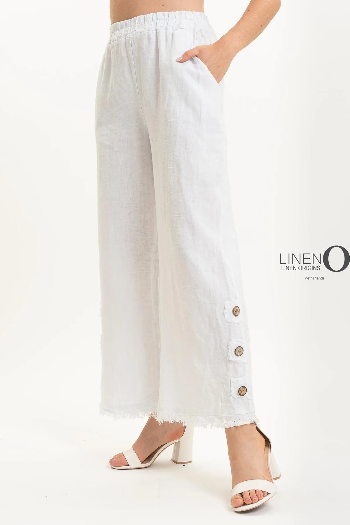 Someone is wearing the French Linen Pant PT839-11 Bianco from LinenO, made of eco-friendly European linen, with wide legs, frayed hems, and three large side buttons. They pair it perfectly with chic white open-toe sandals on a plain white backdrop.