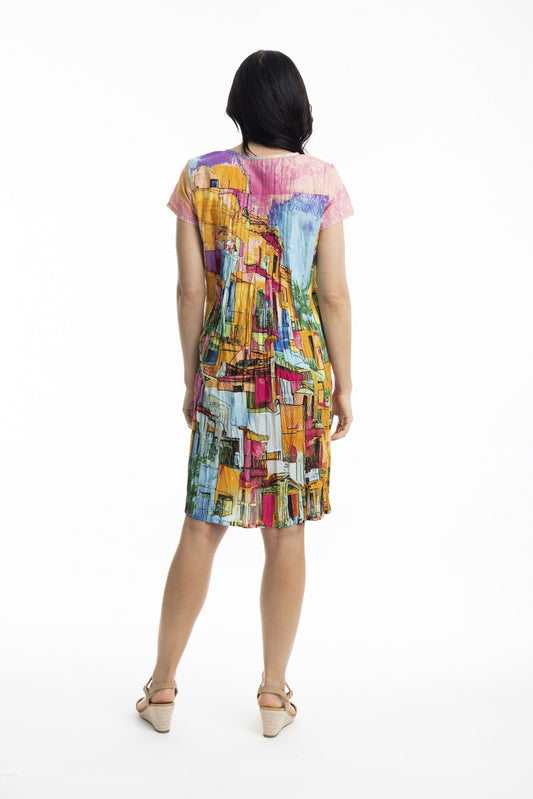 A woman with dark hair, facing away, wears the ORIENTIQUE Printed Cotton Easy Fit Dress Style: 20058 Havana. The knee-length, short-sleeved dress features colourful cityscape-inspired prints and is made from organic cotton, paired with beige sandals on a white background.