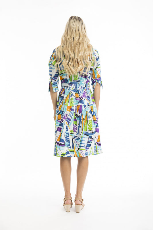 A woman with long wavy blonde hair faces away, wearing the ORIENTIQUE Cannes Collared Dress with Belt (Style: 40031, Print - ORIENTIQUE), featuring a colorful sailboat pattern and A-line silhouette, paired with beige heels against a white background.