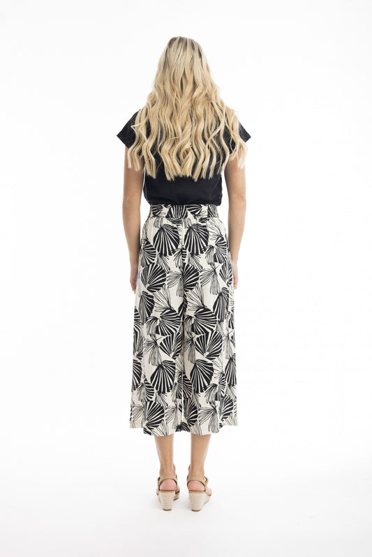 A woman with long, wavy blonde hair stands facing away, wearing ORIENTIQUE's Papagayo Pants Style: 76048—wide-leg linen blend pants with a black and white geometric leaf pattern—plus a black top and light wedge sandals against a white background.