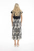A woman with long, wavy blonde hair stands facing away, wearing ORIENTIQUE's Papagayo Pants Style: 76048—wide-leg linen blend pants with a black and white geometric leaf pattern—plus a black top and light wedge sandals against a white background.