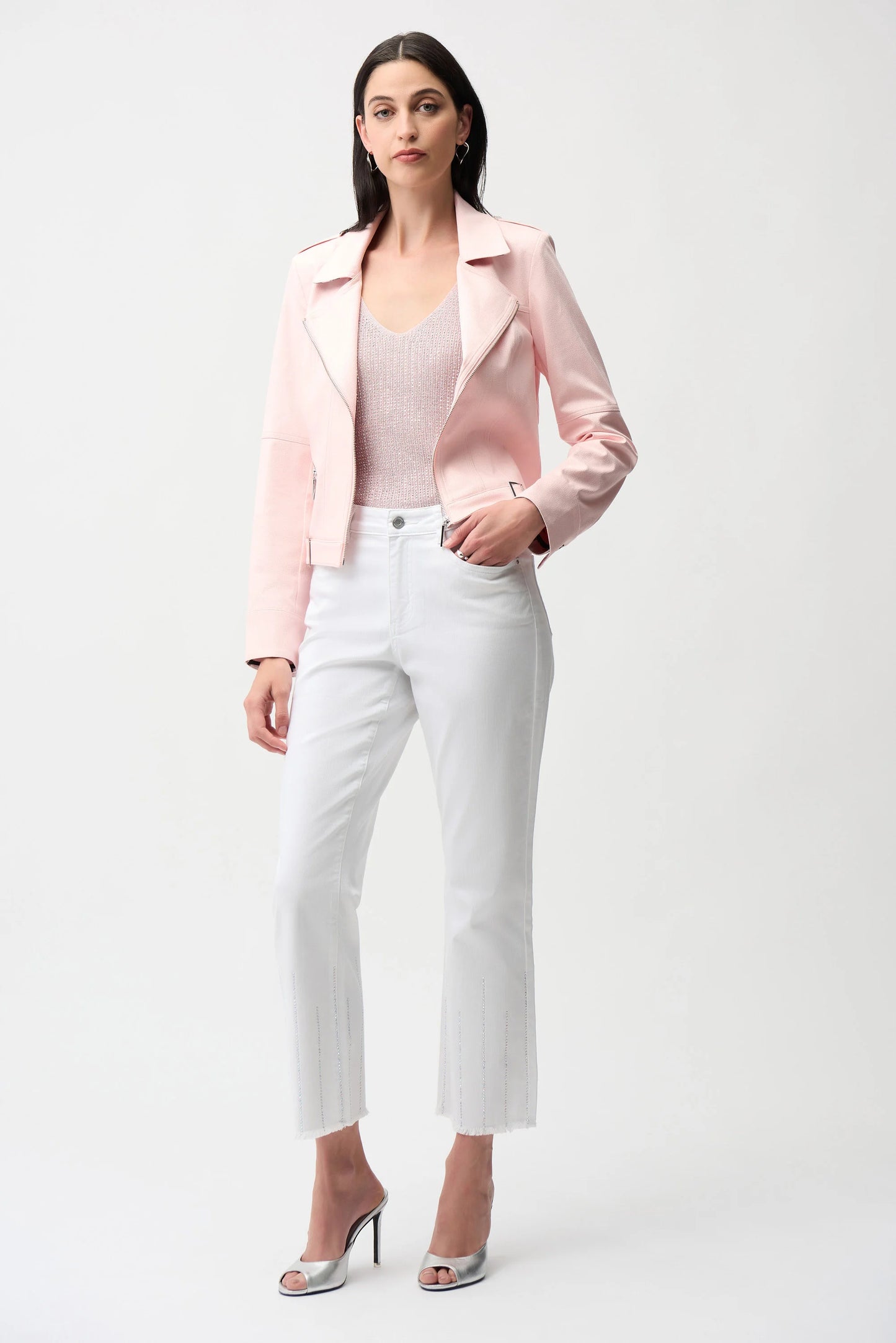 A woman stands against a plain background wearing the Joseph Ribkoff Foiled Faux Suede Fitted Jacket with Decorative Buckles 261910 in light pink, paired with a matching top, white cropped pants, and silver heels. She has straight dark hair and a neutral expression.