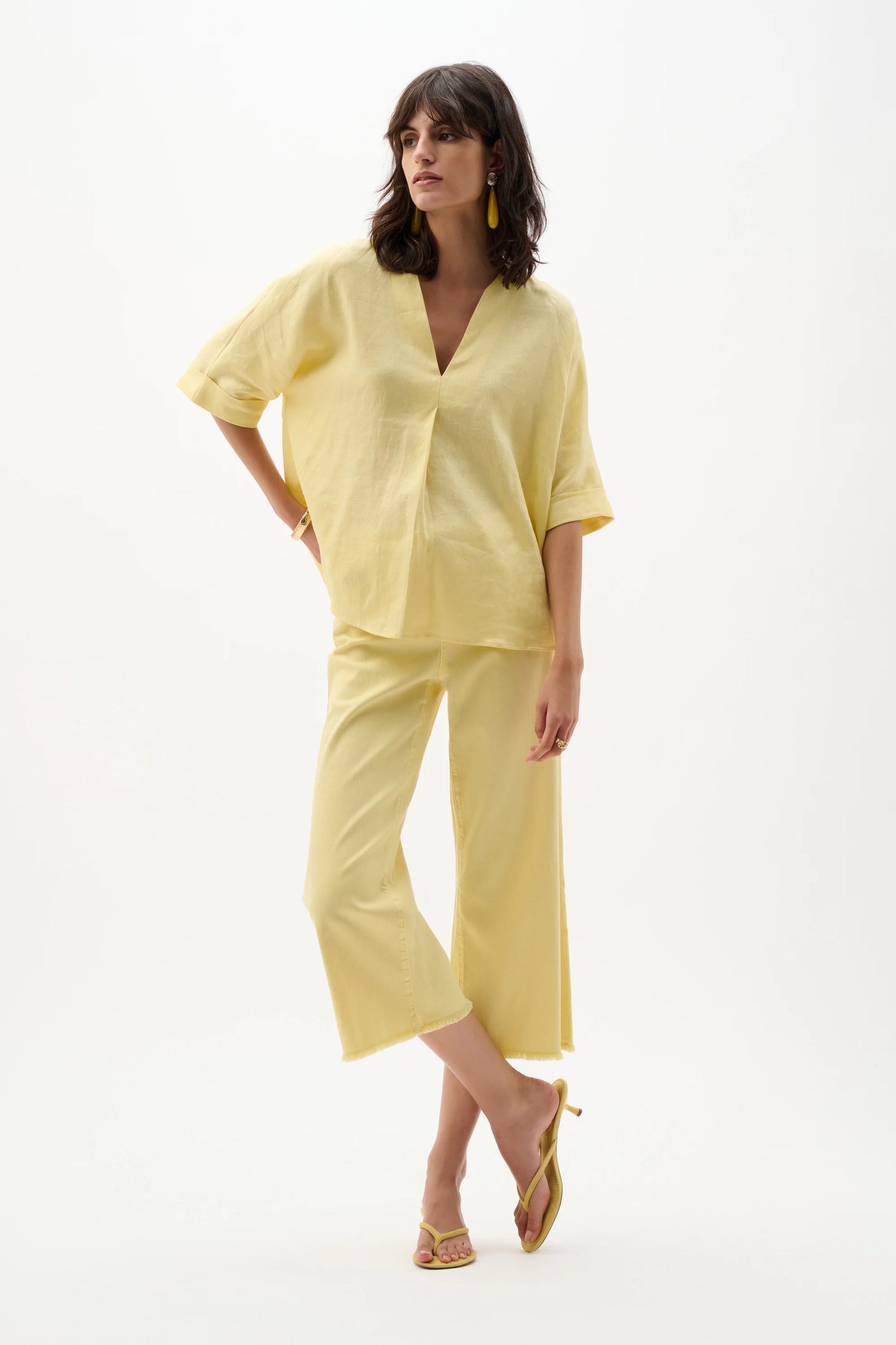A woman poses against a white background wearing the Joseph Ribkoff Linen Straight Top With V-notch neckline in Mellow Yellow, paired with matching wide-legged cropped pants, yellow heeled sandals, and yellow earrings.