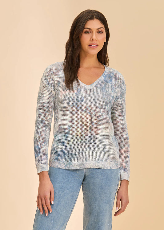 A woman with long brown hair models the French Dressing Jeans V-Neck Printed Sweater in Neutral Narnia, featuring faded floral patterns, paired with light blue jeans and set against a beige background.