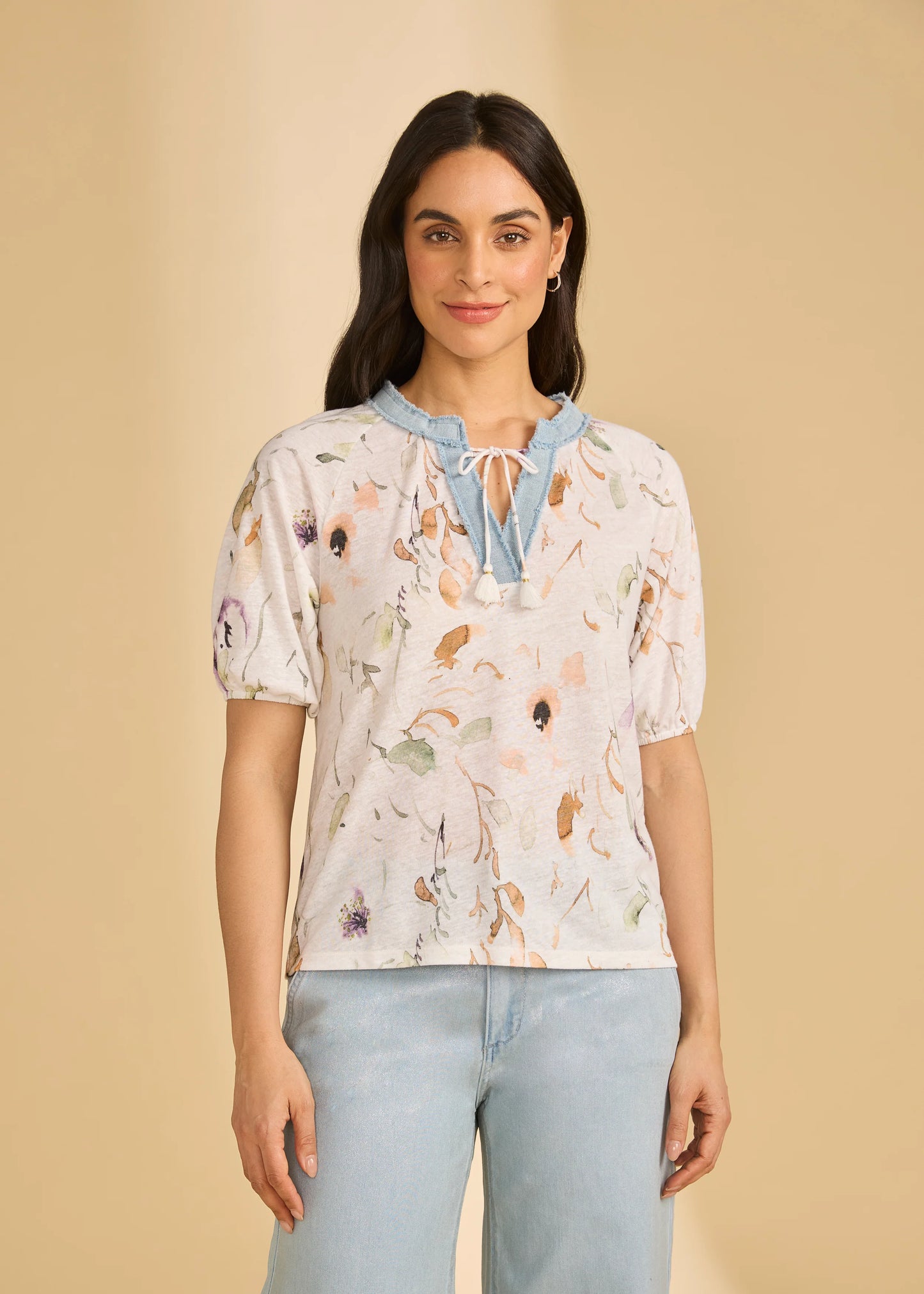 A woman with long dark hair smiles softly in a French Dressing Jeans 3/4 Sleeve Embellished Top (Delicate 3877788-PRT) and light-wash jeans, standing against a beige background.