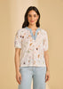 A woman with long dark hair smiles softly in a French Dressing Jeans 3/4 Sleeve Embellished Top (Delicate 3877788-PRT) and light-wash jeans, standing against a beige background.