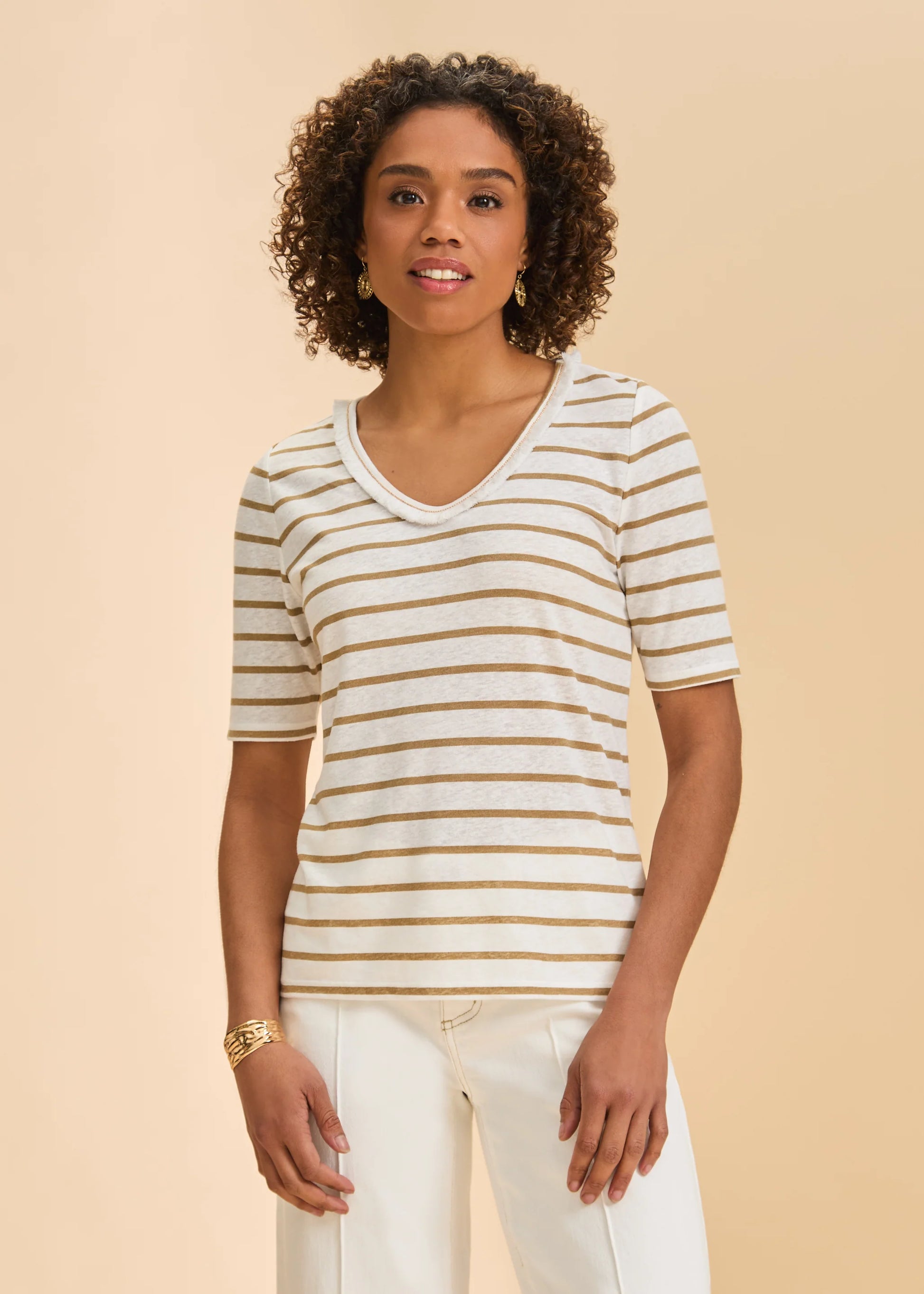 A woman with curly hair, smiling gently at the camera, wears the French Dressing Jeans Elbow Sleeve Fringed Neck Top in henna against a beige background.