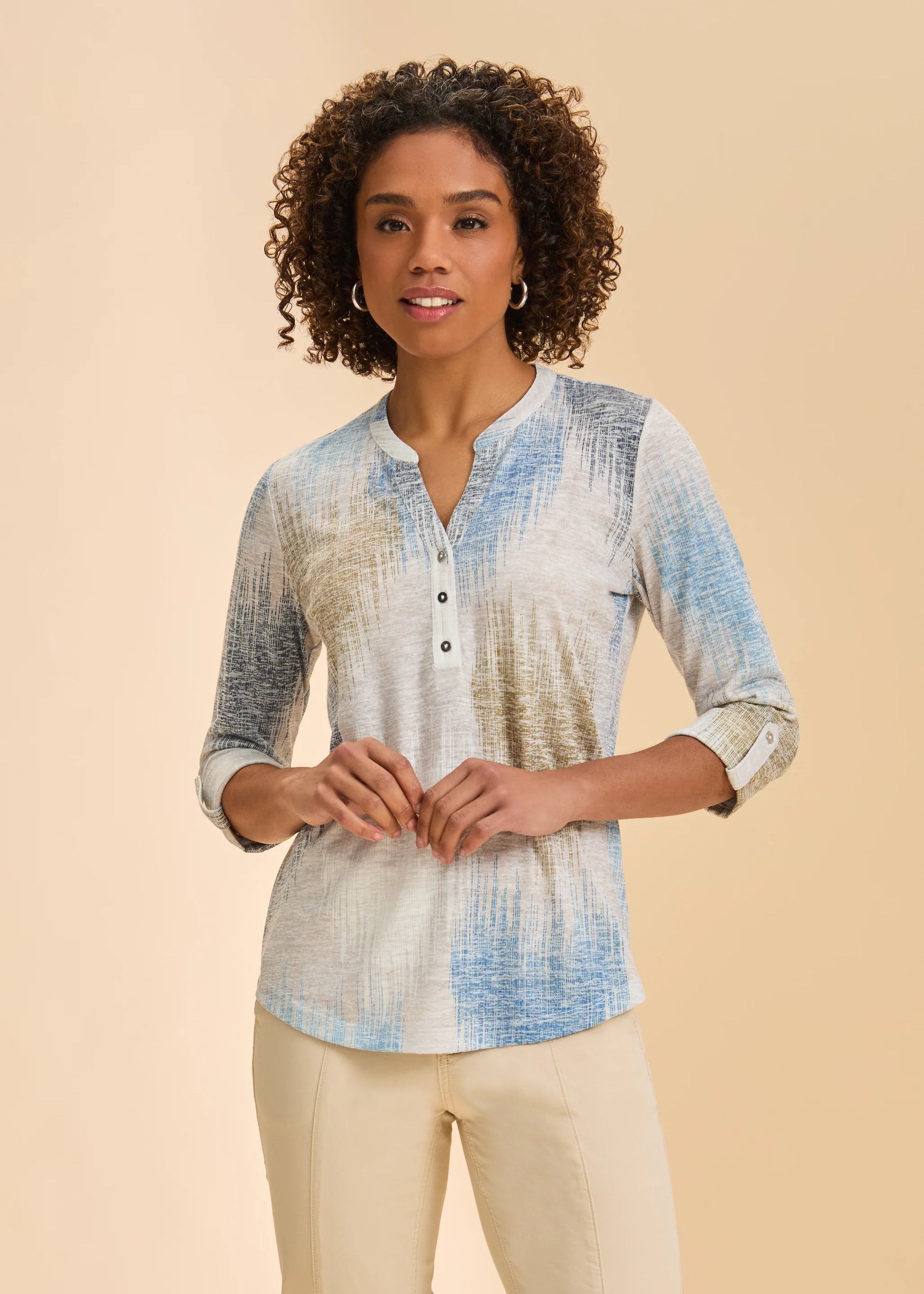 A woman with curly hair smiles gently, wearing the French Dressing Jeans 3/4 Sleeve Henley Top in Neutral Ikat (blue, beige, and gray abstract design) paired with light-colored pants, standing against a plain beige background.