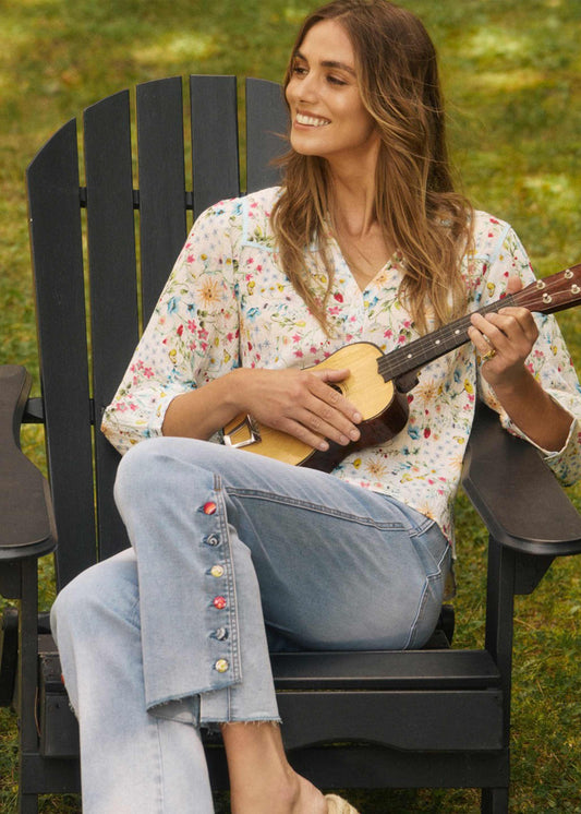 A woman smiles on the grass in a black outdoor chair, playing a small guitar. She wears a floral shirt and French Dressing Jeans Pull-On Boot Crop Denim in Blue Eclipse, featuring colorful buttons and stretchy comfort.