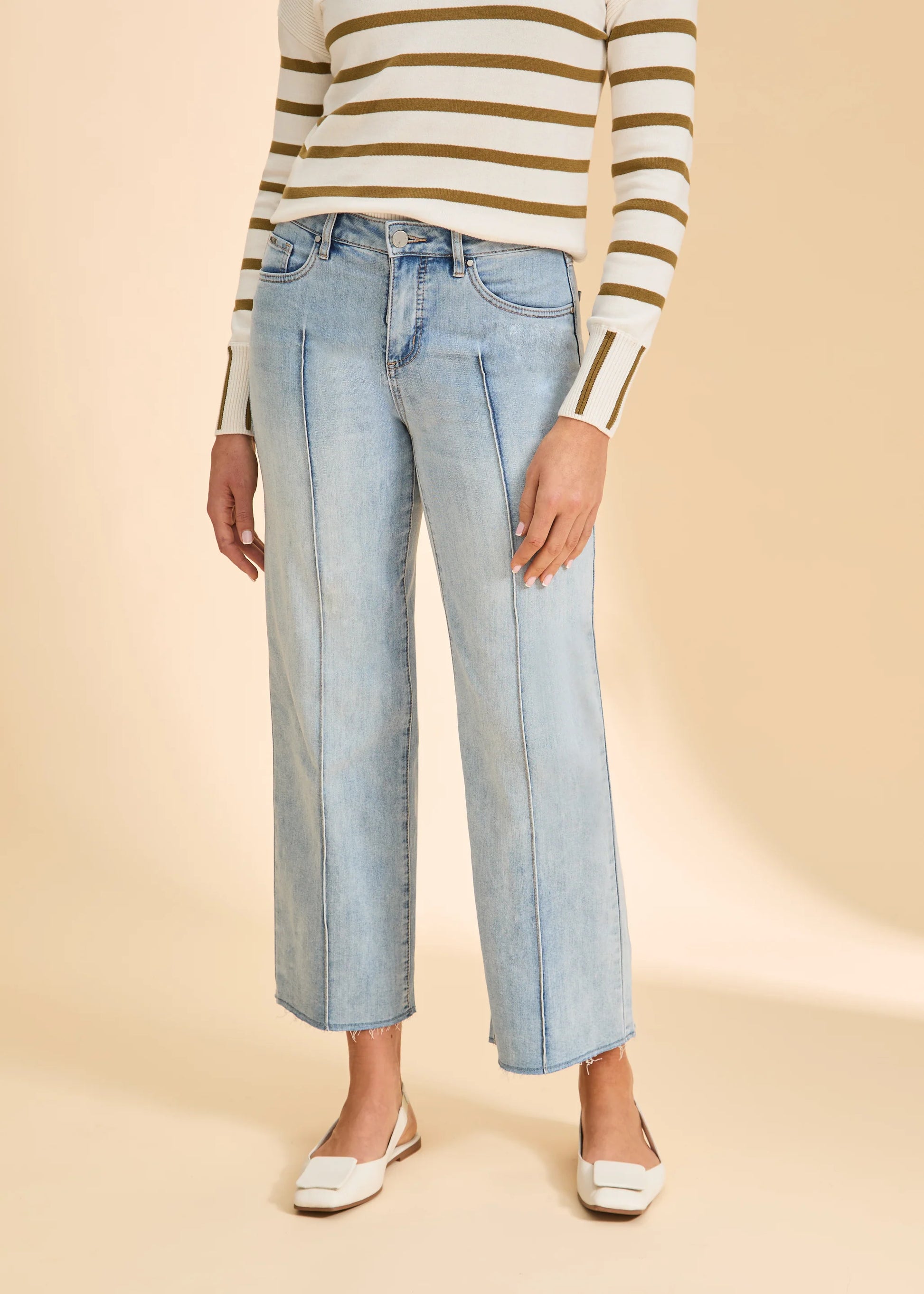 A person models French Dressing Jeans’ Olivia Wide Ankle Denim in Pale Wash, pairing the wide-leg jeans with a long-sleeve white sweater with olive green stripes and white low-heeled shoes, standing against a beige background.