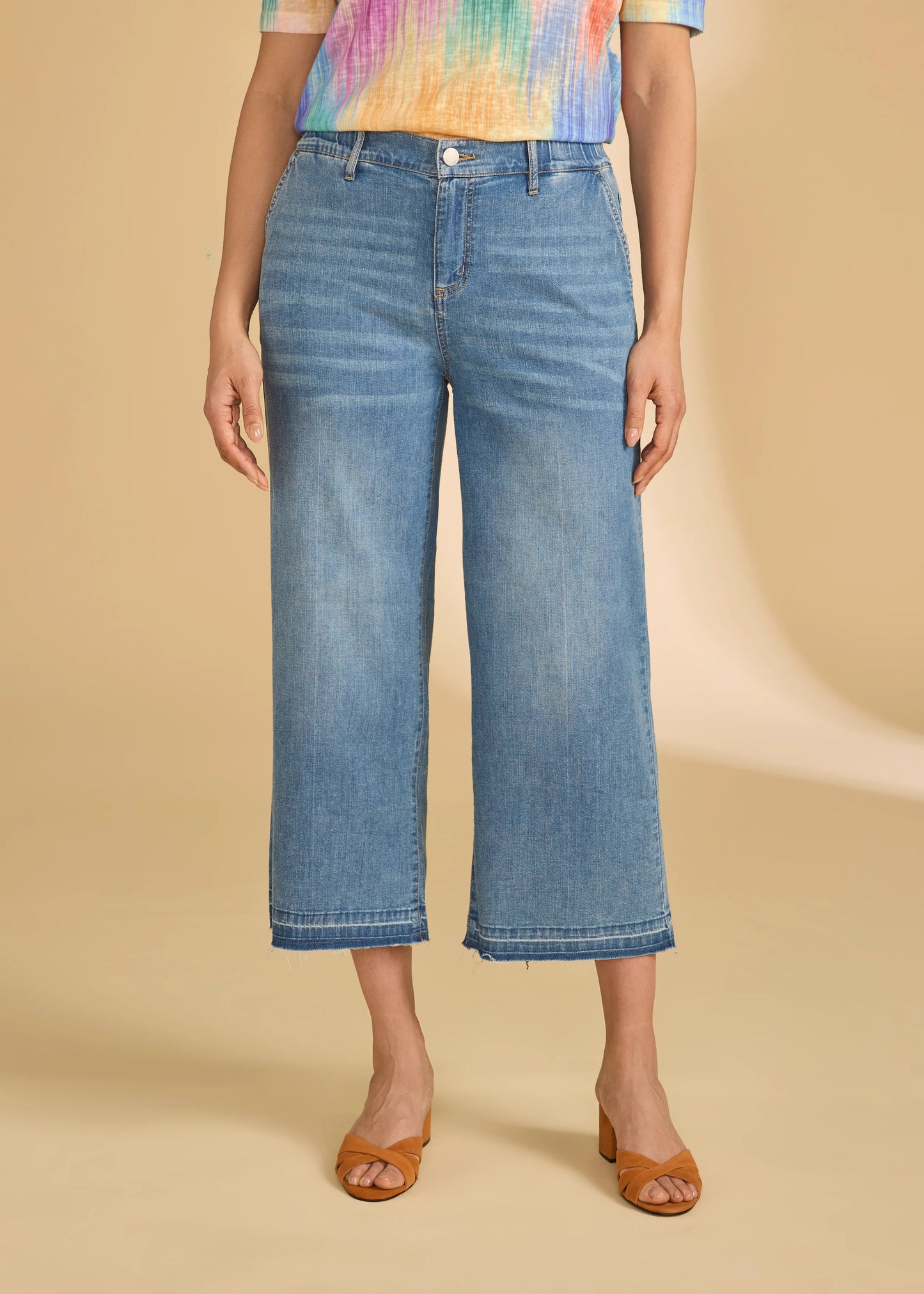 A person wears French Dressing Jeans Pull-On Wide Crop Trouser Denim in Medium Wash with brown open-toe heels and a colorful short-sleeve top, set against a beige background.