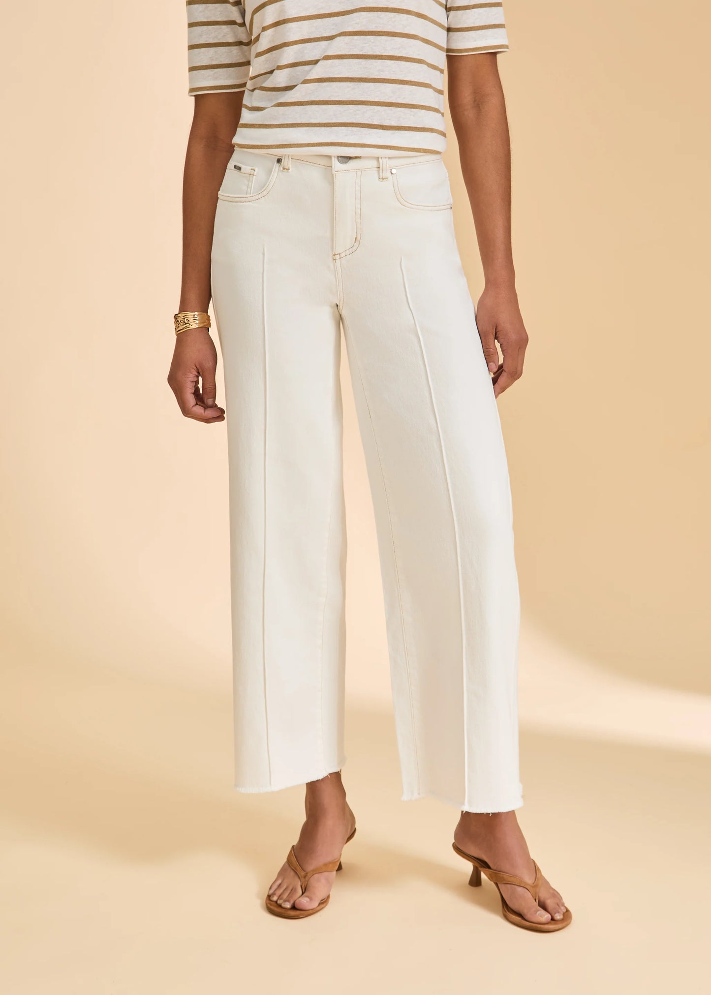 A person wears a beige-and-white striped short-sleeve top, tan heeled sandals, a gold bracelet, and French Dressing Jeans Olivia Wide Leg Ankle Denim in Ivory (2201948-IVR), standing against a beige background.