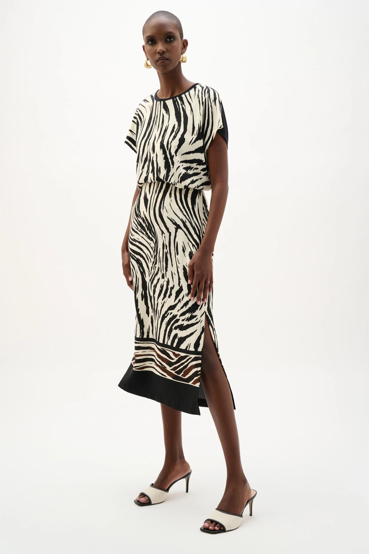 A woman models the Joseph Ribkoff Recycled Woven Animal Print Straight Skirt 262050, styled with black and white heeled sandals and gold hoop earrings, standing against a plain white background.