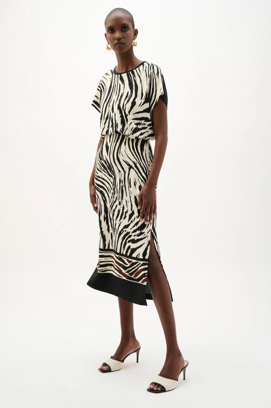 A woman models the Joseph Ribkoff Recycled Woven Animal Print Straight Skirt 262050, styled with black and white heeled sandals and gold hoop earrings, standing against a plain white background.