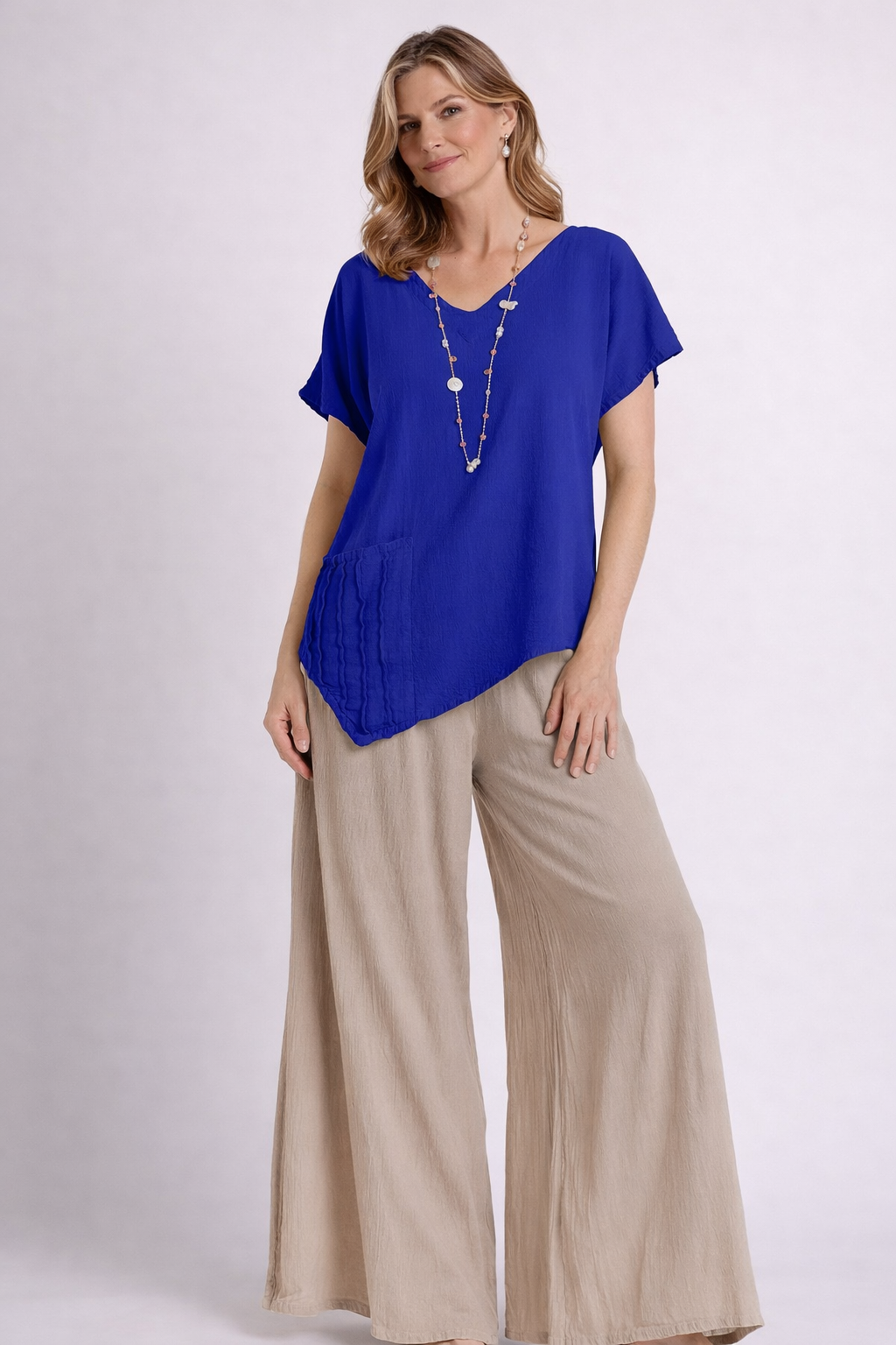 A woman wears the Oh My Gauze Polly Cobalt Top T433, featuring an asymmetrical short sleeve and textured pocket, styled with beige wide-leg pants and a beaded necklace for a light, airy, and breathable summer look.
